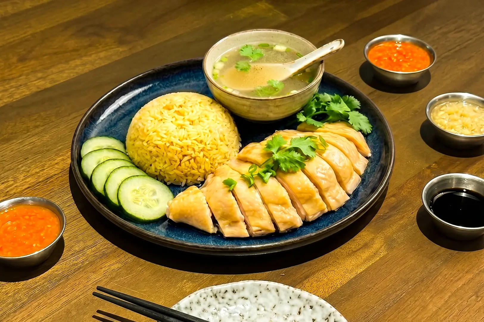 Hainanese chicken rice with sliced poached chicken, turmeric rice, cucumber, dipping sauces, and soup at a trendy Singapore restaurant, captured in angled overhead shot