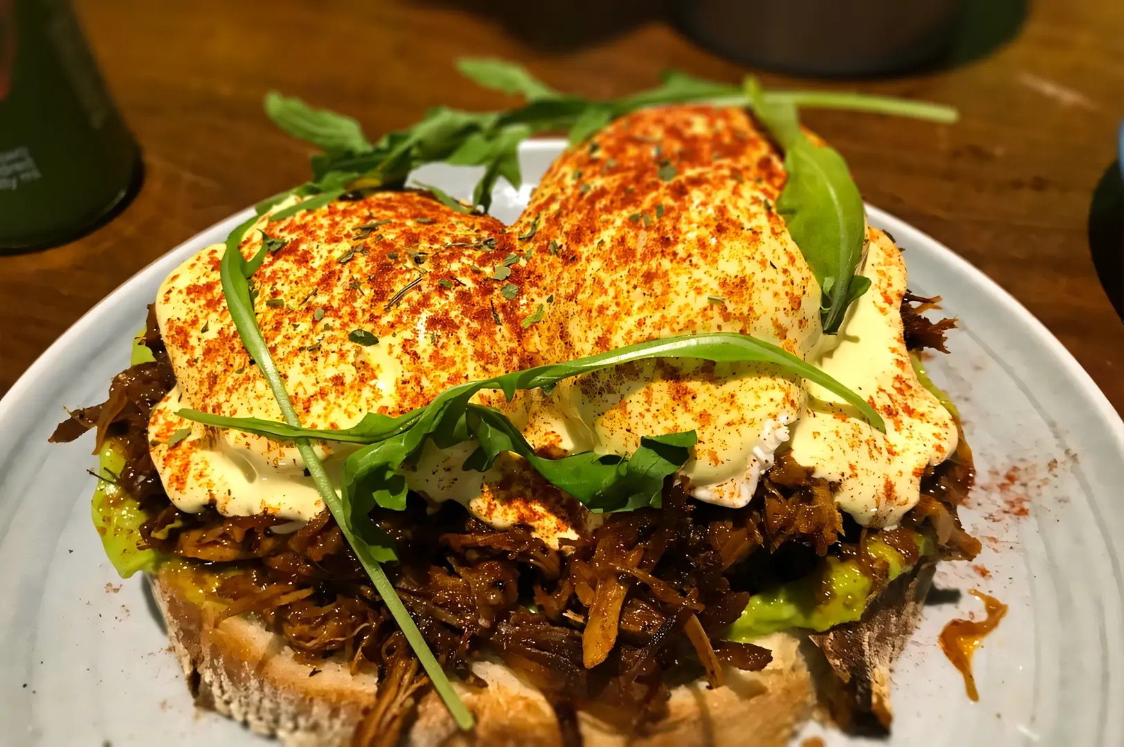 Close-up angle shot of pulled pork and avocado Eggs Benedict on sourdough at Lola’s Cafe Kovan, topped with creamy hollandaise sauce, paprika seasoning, and fresh arugula – popular brunch dish in Singapore