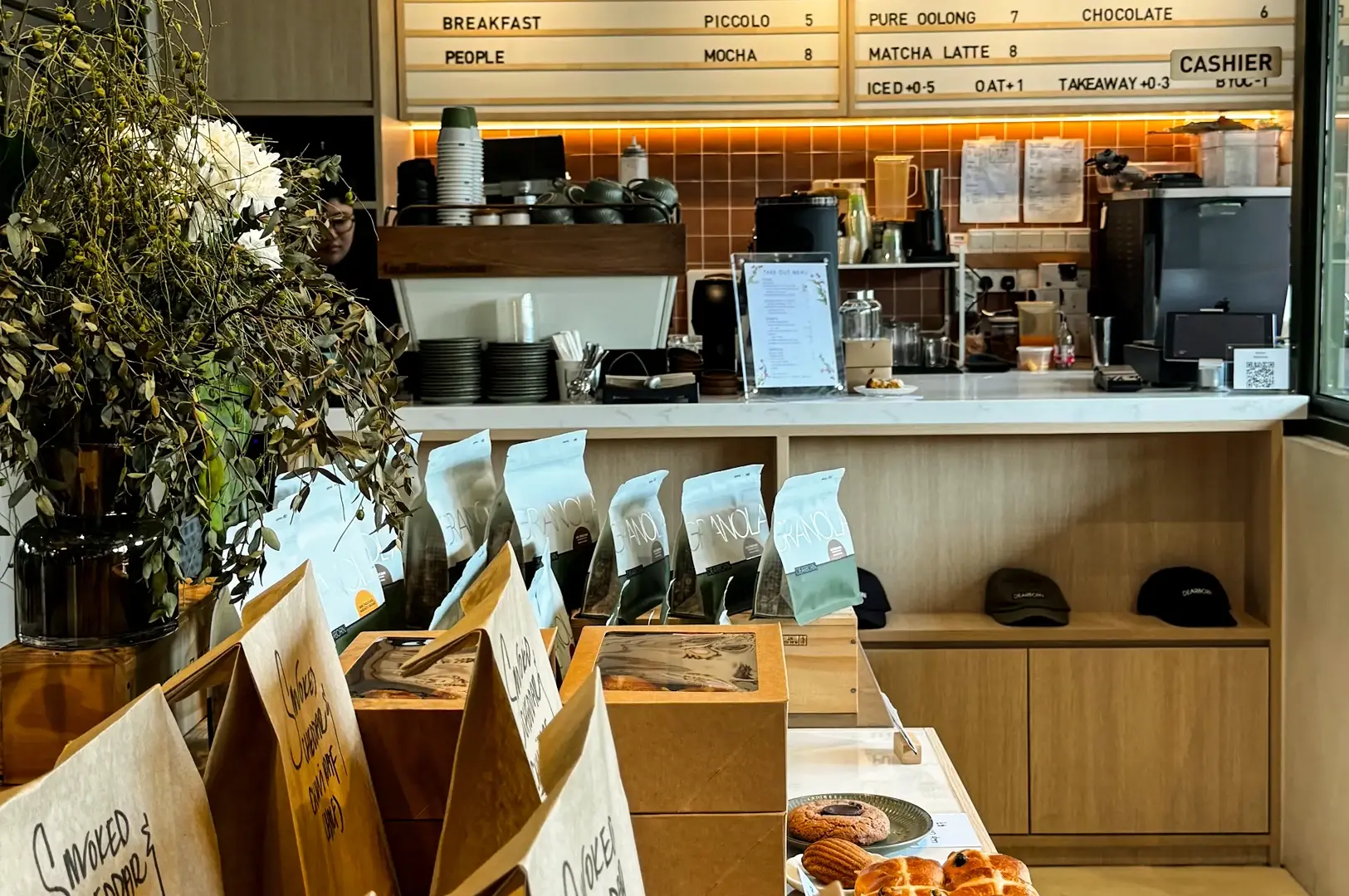 An eye-level, medium interior shot of the ordering and cashier counter at Dearborn Singapore, showcasing a minimalist menu board on the wall, retail bags of house-made granola displayed in the foreground, and fresh baked goods on a platter.