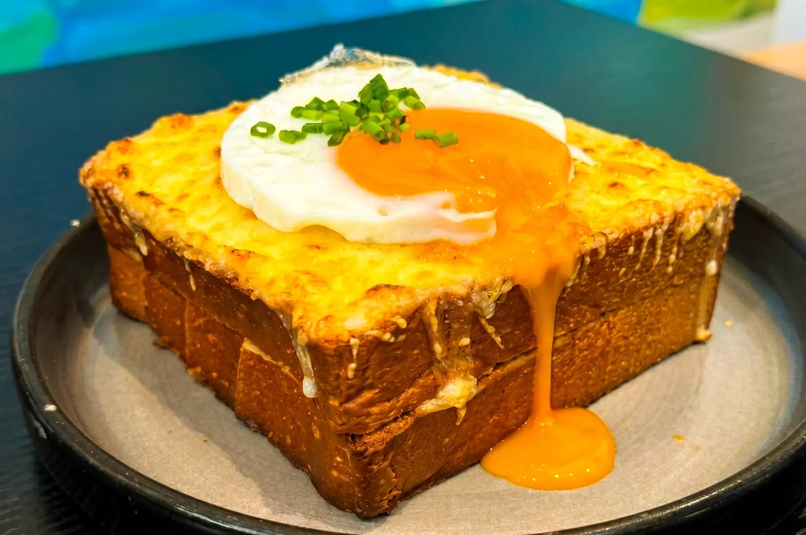 Thick kaya toast with melted cheese and soft poached egg with runny yolk on top, modern Singapore breakfast dish photographed in close-up hero angle