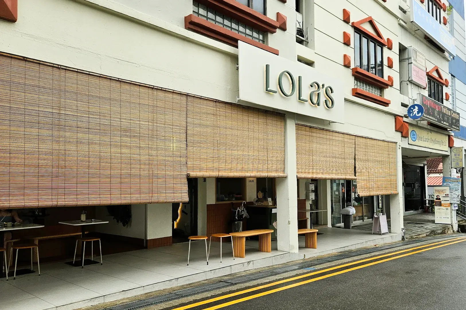 Wide street-level angle shot of Lola’s Cafe Kovan exterior along Simon Road near Kovan MRT, showing minimalist facade, outdoor seating, and popular Singapore brunch destination
