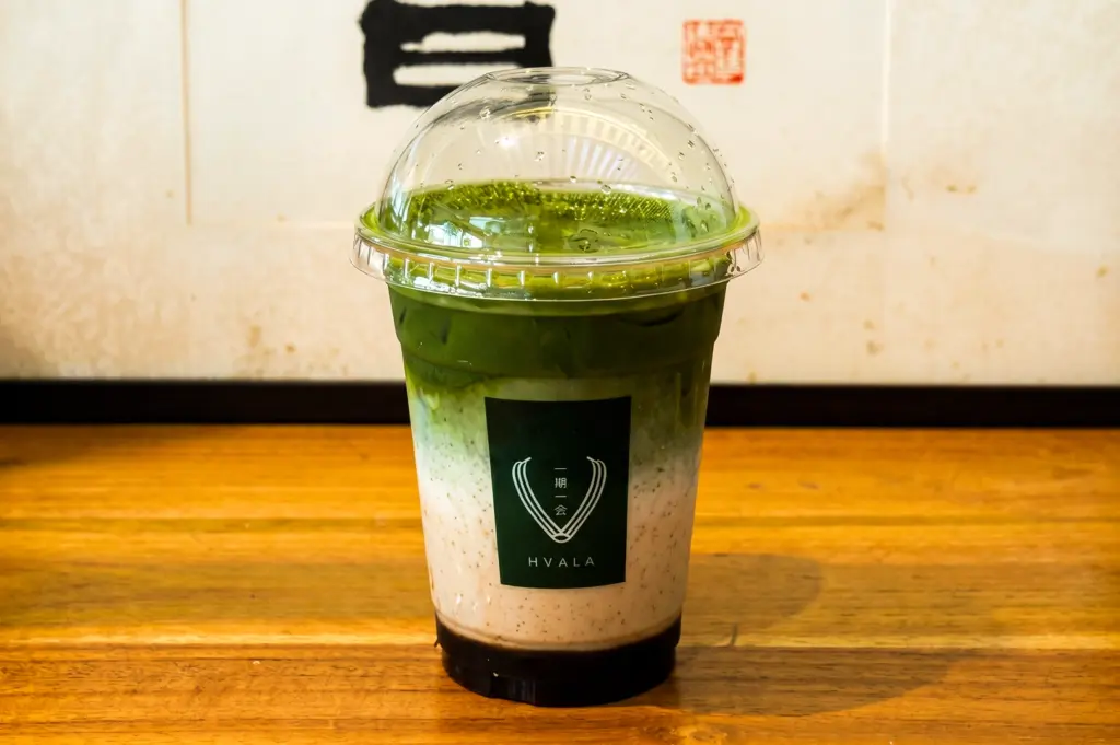 Eye-level shot of layered iced matcha latte in clear cup with dome lid on wooden table at Hvala café, showcasing green matcha over milk and syrup