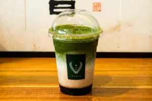 Eye-level shot of layered iced matcha latte in clear cup with dome lid on wooden table at Hvala café, showcasing green matcha over milk and syrup