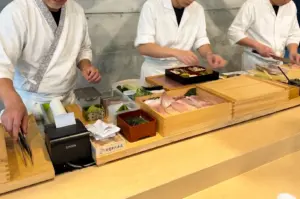 Eye-level wide-angle view of an omakase sushi counter where multiple chefs in white uniforms prepare seafood and ingredients behind a light wood counter. Open wooden trays display assorted fish cuts, garnishes, seaweed, and preparation items, while containers, utensils, and a receipt printer are arranged across the workstation. The background features a textured concrete wall and an organized preparation area, with bright indoor lighting highlighting the natural wood surfaces, fresh ingredients, and craftsmanship of the sushi-making process.