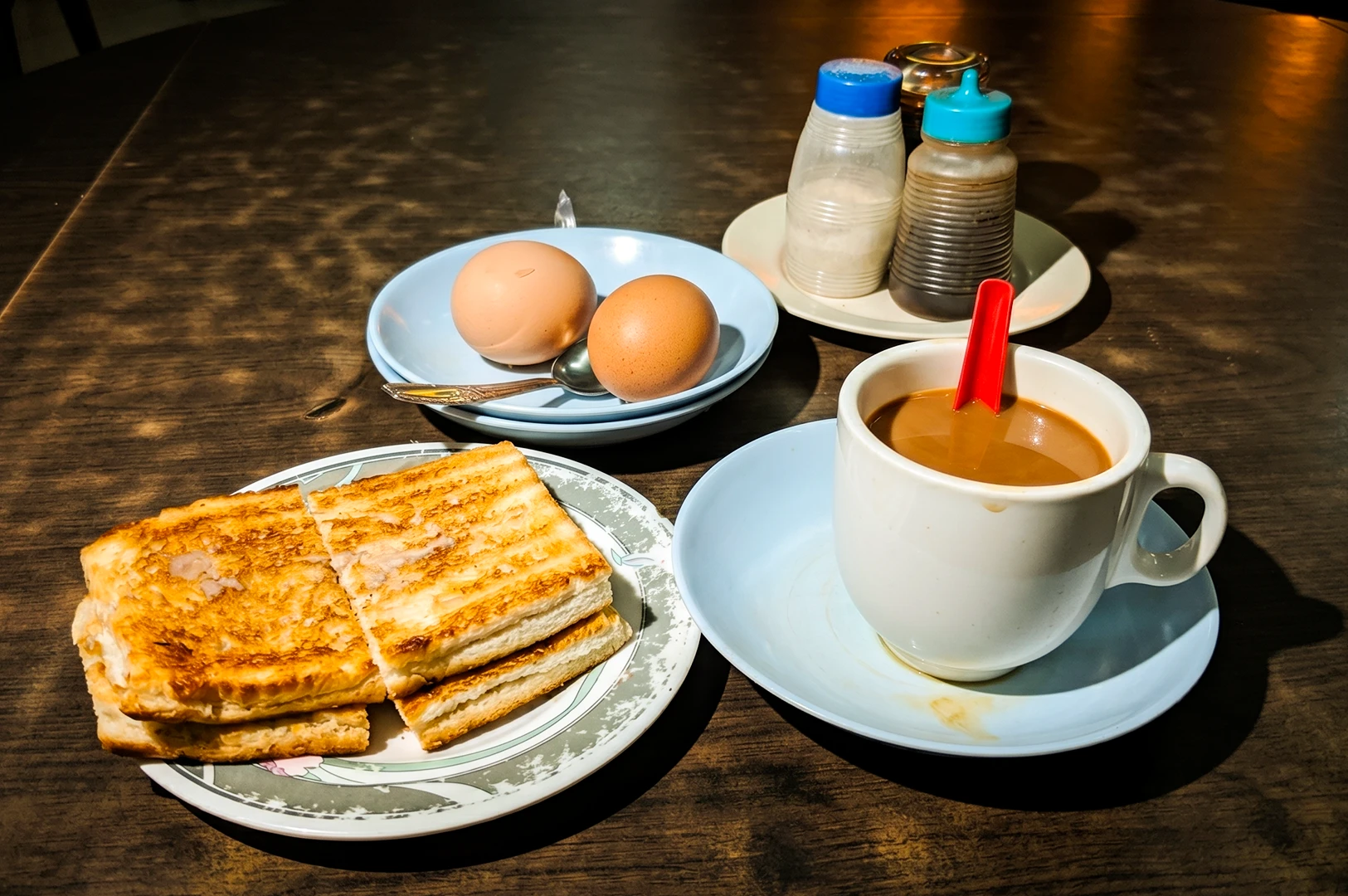 High-angle close-up of kaya toast, soft-boiled eggs, and kopi on a wooden table in a casual kopitiam breakfast setting