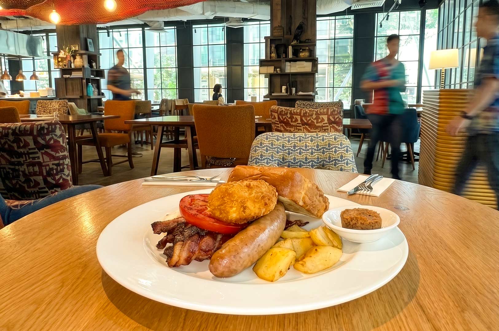 A hearty breakfast platter on a white plate featuring a sausage, bacon, a sliced tomato, a fried hash brown or croquette, roasted potato wedges, a piece of bread, and a small dish of spread, set on a wooden table in a restaurant with large windows and blurred figures in the background.