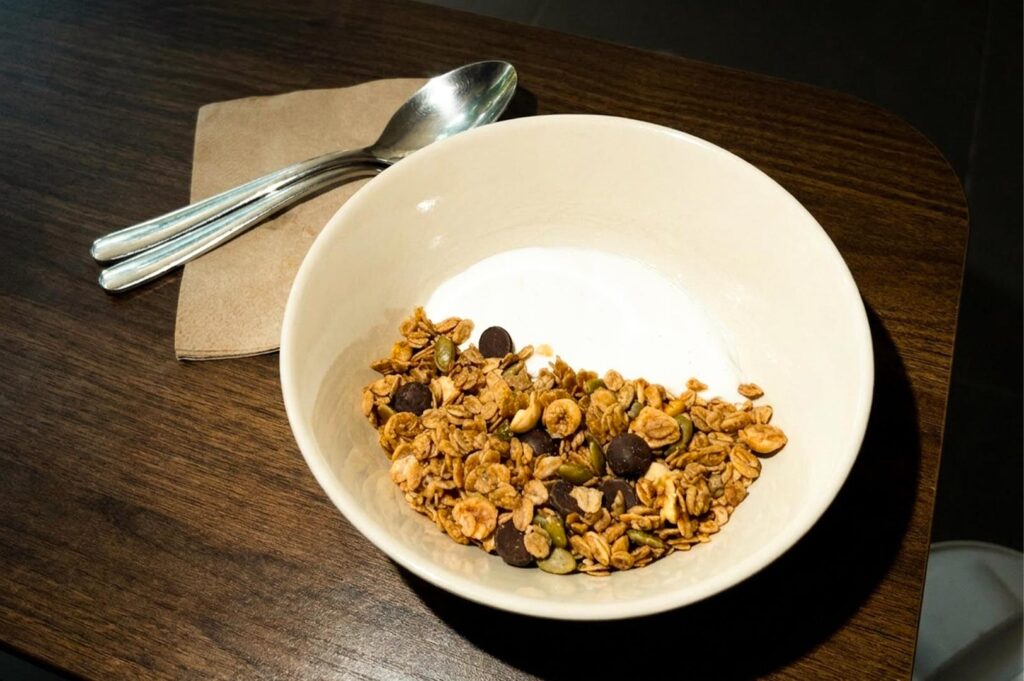 A white ceramic bowl filled with creamy white yogurt topped with granola mix, pumpkin seeds, and chocolate chips, served with a spoon resting on a brown paper napkin on a dark wooden table.