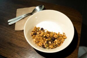 A white ceramic bowl filled with creamy white yogurt topped with granola mix, pumpkin seeds, and chocolate chips, served with a spoon resting on a brown paper napkin on a dark wooden table.