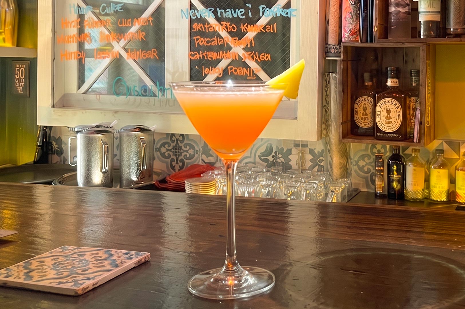 A vibrant orange cocktail in a stemmed martini glass sits on a polished wooden bar counter, garnished with a piece of citrus peel on the rim. In the background, a colorful bar setup features a chalkboard menu with handwritten text, shelves with bottles, a decorative pineapple shaker, and various stickers and polaroids on the wall.