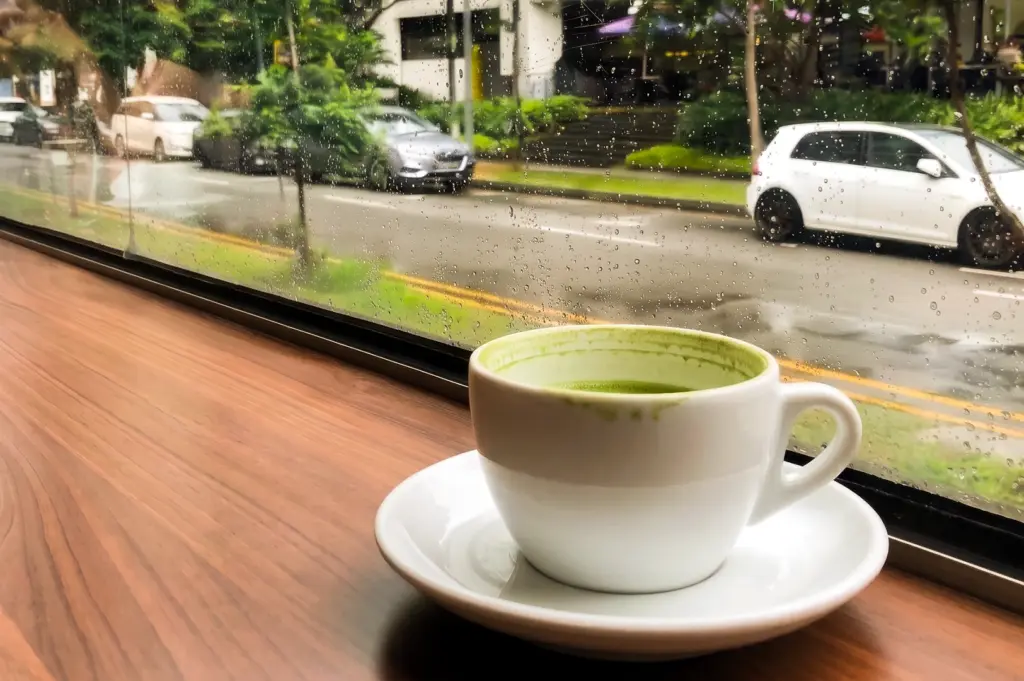 Eye-level close-up shot of a pistachio latte on a café windowside table overlooking a rainy Singapore street, reflecting modern café culture, trendy food habits, and the experience of ordering popular drinks for social appeal.
