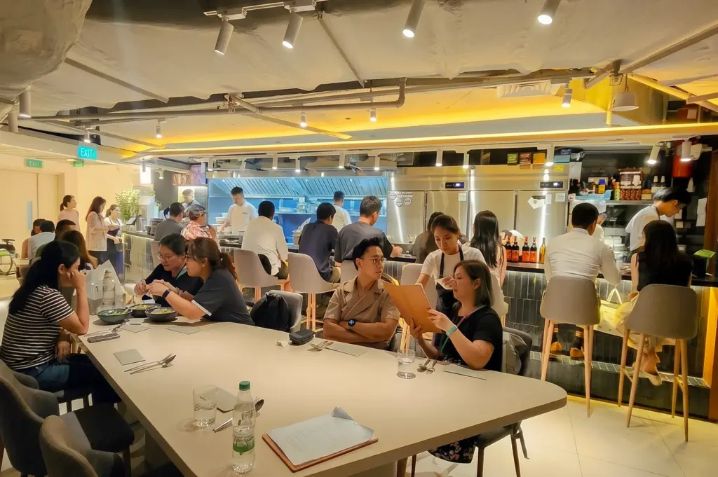 Wide-angle interior view of Sio Pasta Singapore at Raffles City, featuring an open kitchen, counter seating, communal dining table, and guests enjoying Japanese-style handmade pasta.