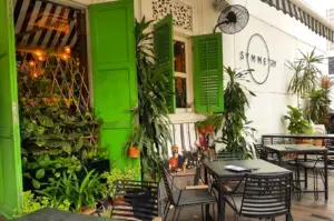Eye-level exterior shot of Symmetry Cafe on Jalan Kubor, Singapore, showcasing green shutters, outdoor seating, lush plants, and the popular brunch café's welcoming entrance near Bugis MRT Station.