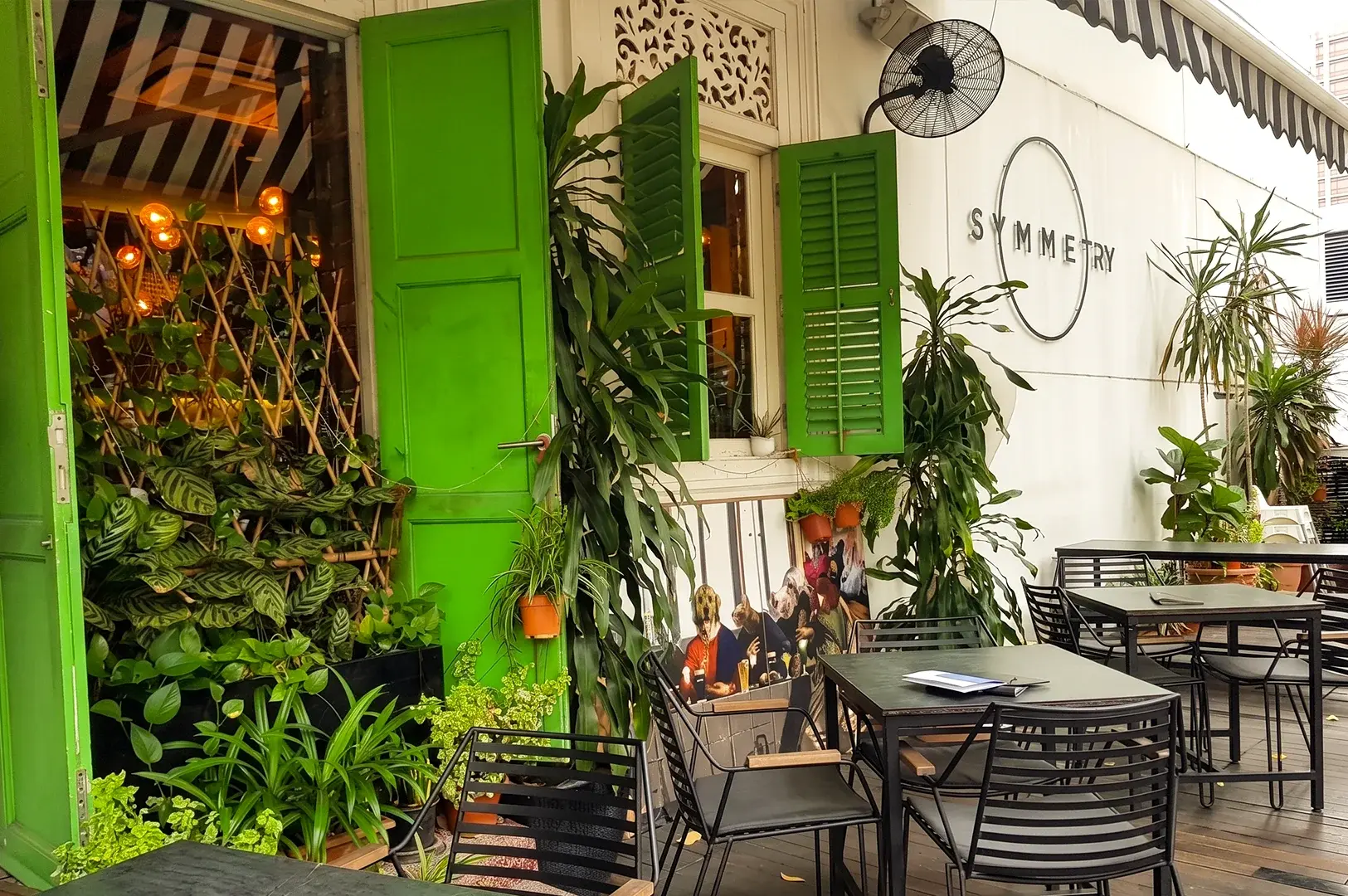 Eye-level exterior shot of Symmetry Cafe on Jalan Kubor, Singapore, showcasing green shutters, outdoor seating, lush plants, and the popular brunch café's welcoming entrance near Bugis MRT Station.