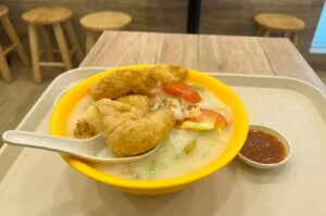 A bright yellow bowl filled with milky fried fish soup, crispy fried fish nuggets, tomato slices, and vegetables, served with a white spoon and a small dish of chili sauce on a dining tray.