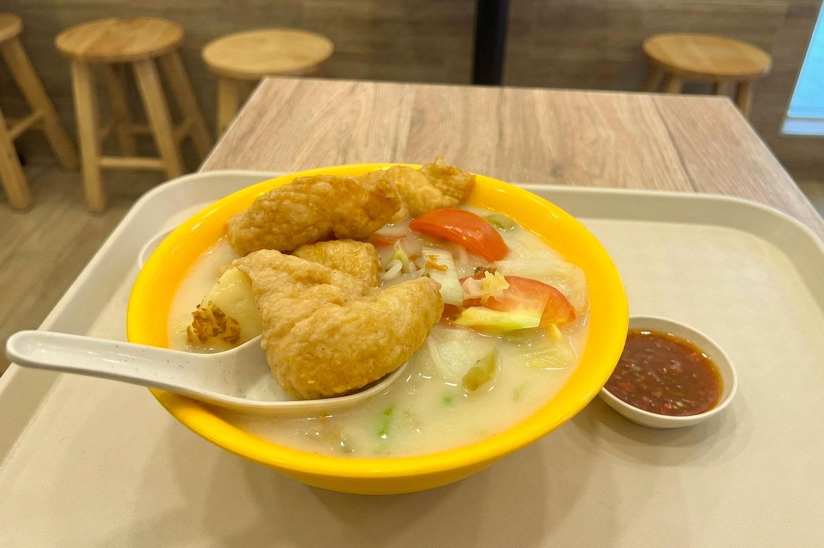 A bright yellow bowl filled with milky fried fish soup, crispy fried fish nuggets, tomato slices, and vegetables, served with a white spoon and a small dish of chili sauce on a dining tray.