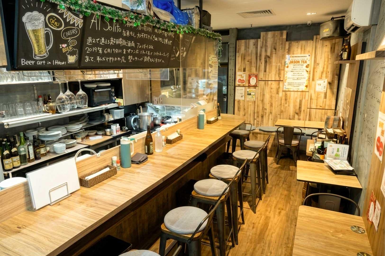 The interior of a cozy Japanese izakaya features a long wooden bar counter lined with high metal stools cushioned in light gray fabric, facing an open kitchen station with hanging wine glasses and shelves of bottles. Above the kitchen, a black chalkboard displays hand-written Japanese text and beer illustrations, while additional wooden two-seater dining tables line the warm, wood-paneled walls in the background.