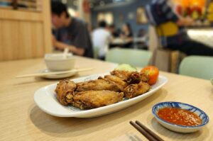 A white oval platter resting on a light wooden table holds several golden-brown fried chicken wings garnishing cucumber slices and a halved tomato. Next to the platter, a small blue-patterned dish filled with chili sauce sits beside a pair of wooden chopsticks, while patrons sit softly blurred in the background of the restaurant dining room.