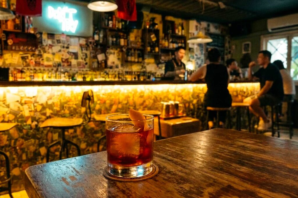 This atmospheric close-up shot focuses on an amber-colored cocktail served on the rocks with a citrus twist garnish, set on a coaster upon a dark, rustic wooden table. In the softly blurred background, the warm, golden glow of a lively dimly lit bar illuminates patrons seated on high stools along the long counter, with a bartender at work in front of shelves filled with spirits and a light-blue neon sign mounted on the wall.