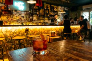 This atmospheric close-up shot focuses on an amber-colored cocktail served on the rocks with a citrus twist garnish, set on a coaster upon a dark, rustic wooden table. In the softly blurred background, the warm, golden glow of a lively dimly lit bar illuminates patrons seated on high stools along the long counter, with a bartender at work in front of shelves filled with spirits and a light-blue neon sign mounted on the wall.
