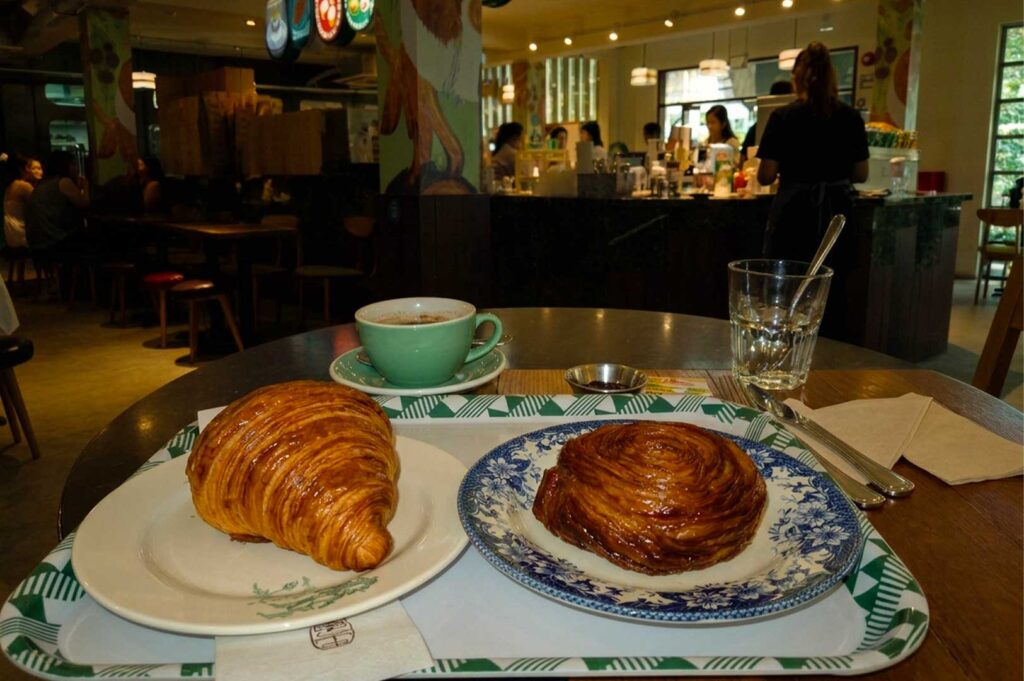 A table-level shot featuring fresh bakery items set on a tray with green geometric pattern borders. In the foreground, a golden-brown butter croissant sits on a light ceramic plate, placed next to a blue-patterned vintage plate holding a round, golden swirl pastry. Behind the pastries rests a light green ceramic teacup filled with hot coffee on its saucer, accompanied by a glass of water with a spoon. The background shows a warmly lit cafe interior with staff working behind an ordering counter.