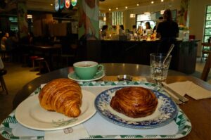 A table-level shot featuring fresh bakery items set on a tray with green geometric pattern borders. In the foreground, a golden-brown butter croissant sits on a light ceramic plate, placed next to a blue-patterned vintage plate holding a round, golden swirl pastry. Behind the pastries rests a light green ceramic teacup filled with hot coffee on its saucer, accompanied by a glass of water with a spoon. The background shows a warmly lit cafe interior with staff working behind an ordering counter.
