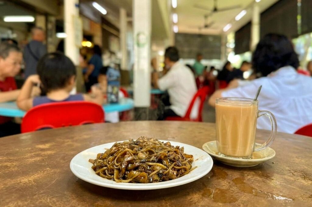 A plate of dark stir-fried flat noodles (Hokkien mee) served on a white plate alongside a clear glass mug of hot milk tea (teh tarik) resting on a saucer. The meal sits on a round wooden table, with a bustling open-air kopitiam or hawker hall filled with patrons sitting in red plastic chairs visible in the soft-focus background.