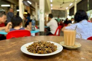 A plate of dark stir-fried flat noodles (Hokkien mee) served on a white plate alongside a clear glass mug of hot milk tea (teh tarik) resting on a saucer. The meal sits on a round wooden table, with a bustling open-air kopitiam or hawker hall filled with patrons sitting in red plastic chairs visible in the soft-focus background.