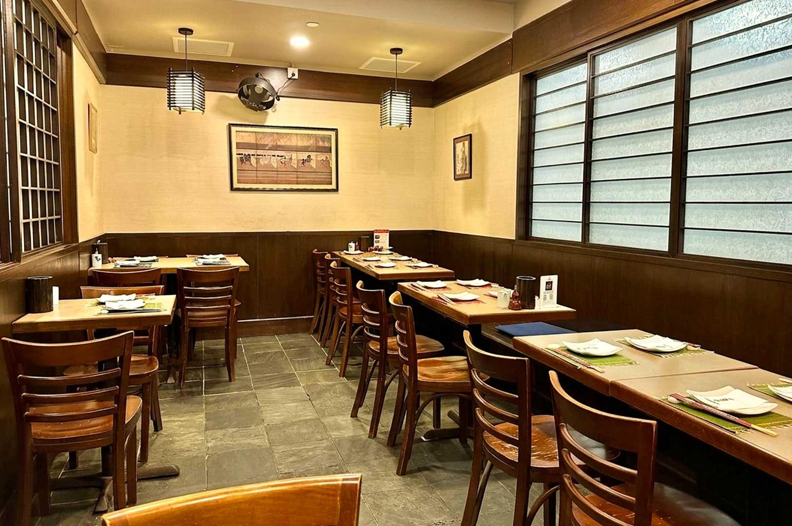 An empty dining area featuring several small wooden tables neatly set for service with green placemats, white plates, and chopsticks. Background: The room features traditional Japanese decor with dark wood wainscoting, pale textured walls, framed artwork, hanging square lanterns, and a large frosted glass window with wooden slats over a dark slate-tiled floor.