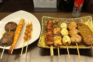 A white curved plate on the left holding grilled shiitake mushroom skewers, a large grilled prawn, and ginkgo nut skewers. To the right, a textured rectangular ceramic plate features assorted yakitori skewers including glazed meat, chicken and leek (negima), quail eggs, minced chicken meatballs (tsukune), and sliced beef.