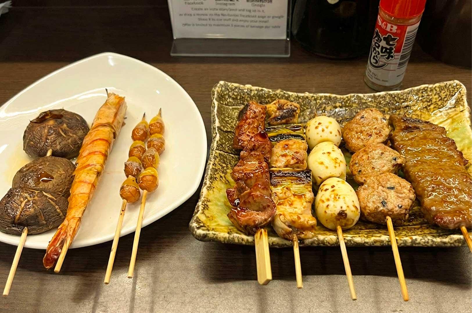 A white curved plate on the left holding grilled shiitake mushroom skewers, a large grilled prawn, and ginkgo nut skewers. To the right, a textured rectangular ceramic plate features assorted yakitori skewers including glazed meat, chicken and leek (negima), quail eggs, minced chicken meatballs (tsukune), and sliced beef.
