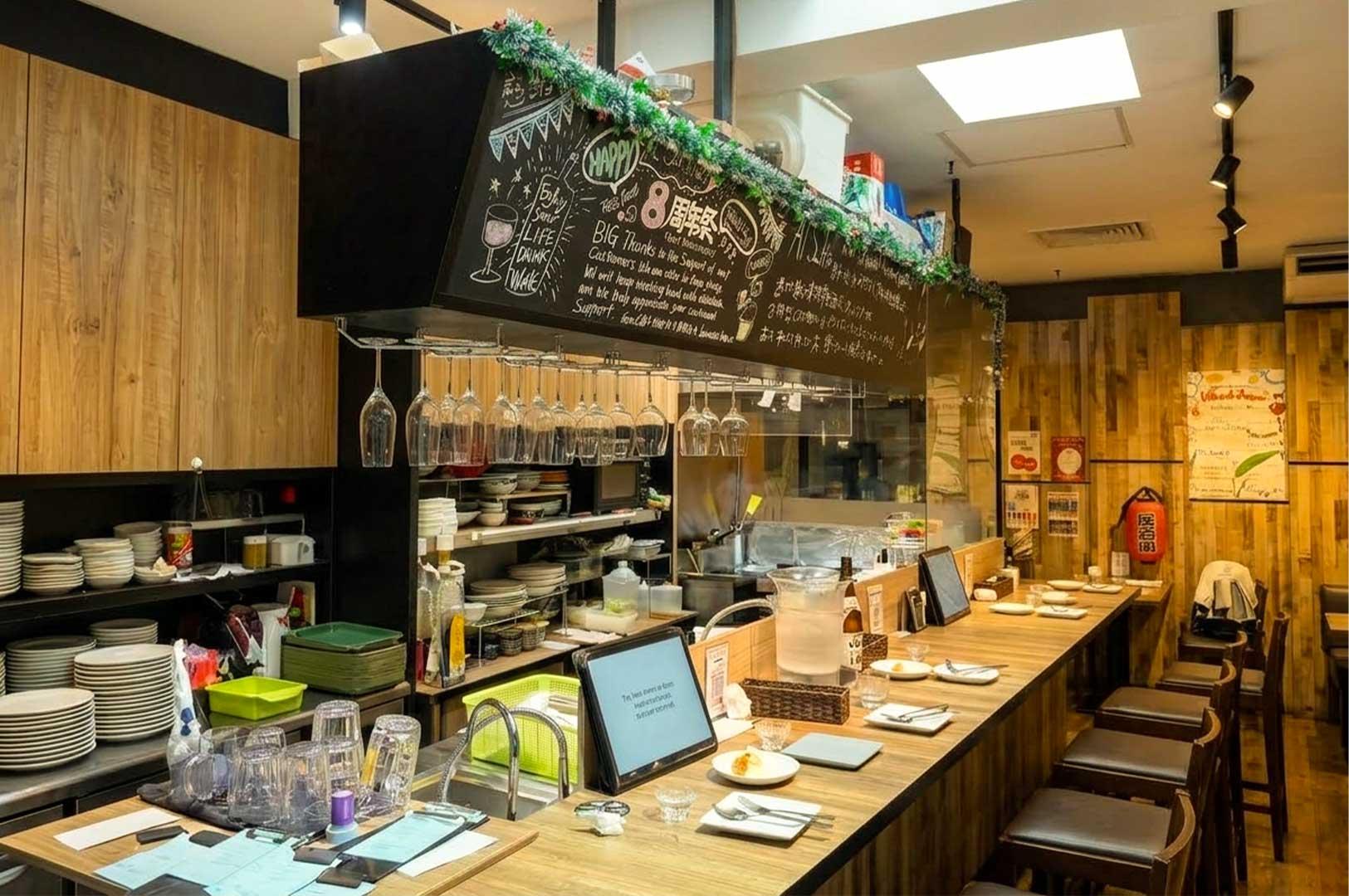 A high-angle view of a Japanese restaurant counter reveals a long wooden bar lined with leather-padded high stools, fully set with plates, cutlery, and drinking glasses for guests. Above the open service area, a black overhead structure decorated with handwritten chalk drawings and festive greenery holds hanging wine glasses, while stacks of clean white dishes and kitchen prep counters sit neatly organized on the left under warm wooden paneling.