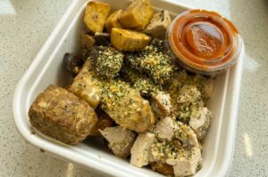 A white square takeout container filled with a healthy protein bowl featuring cubed tempeh, seasoned diced chicken breast sprinkled with herbs, roasted mushrooms, and cubed tofu.