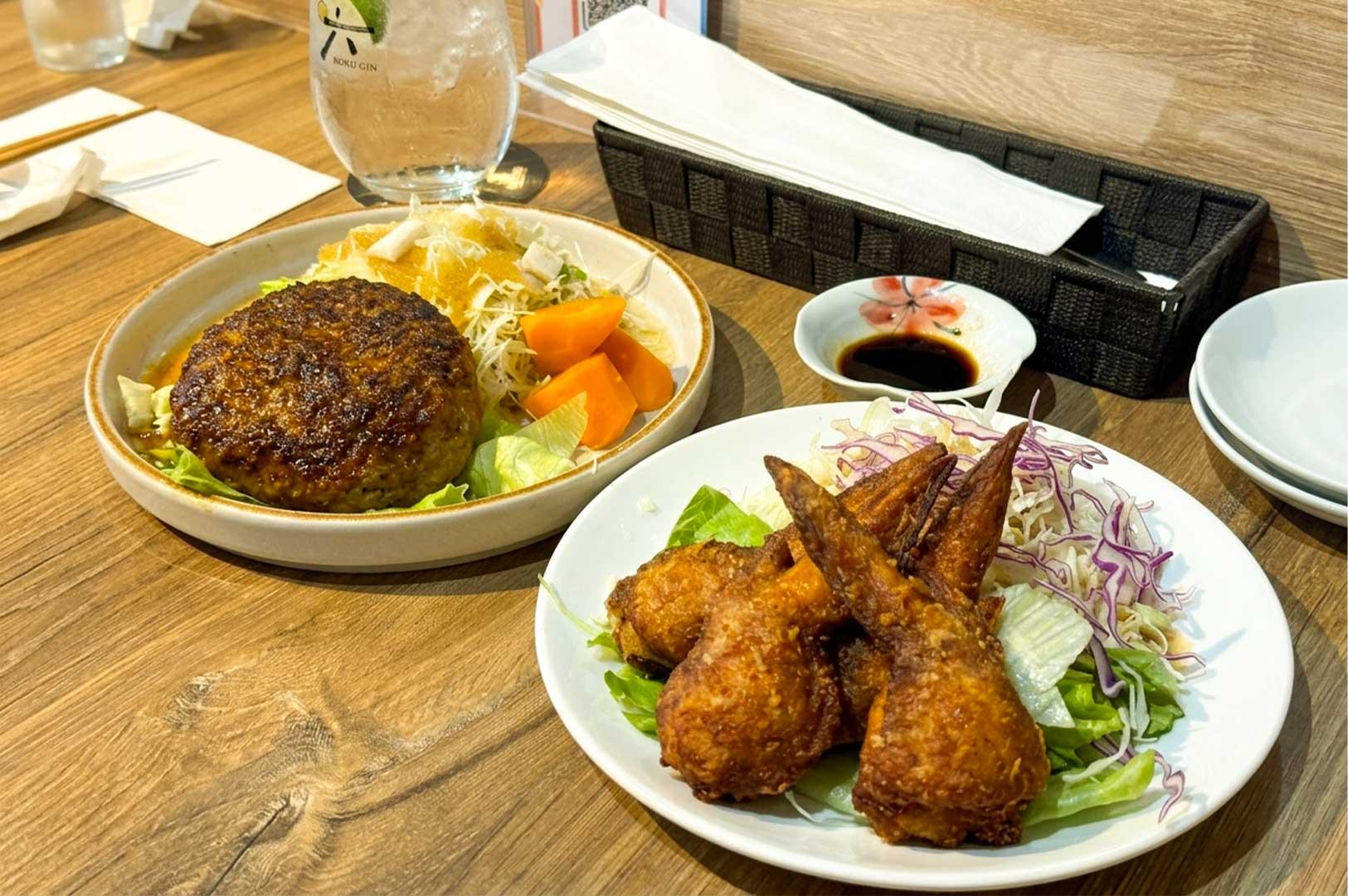 A wooden table display features two Japanese dishes, including a thick hambagu steak served with shredded cabbage and steamed carrots on the left, alongside a plate of golden-brown fried chicken wings paired with a shredded cabbage and purple cabbage salad on the right. In the background, an iced beverage glass, a small bowl of dark dipping sauce, a woven napkin basket, and extra side plates complete the dining setting.