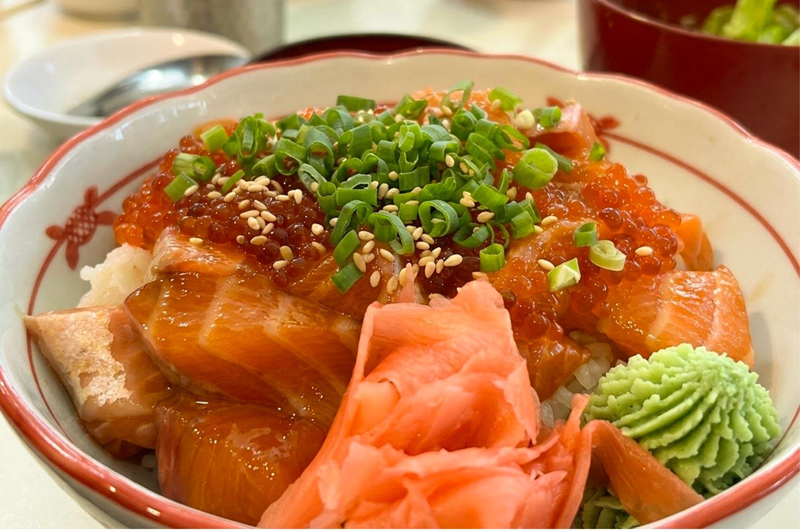 A close-up view of a salmon chirashi don served in a white bowl with a scalloped red rim. The rice is topped with thick, glossy slices of fresh raw salmon and glistening salmon roe (ikura), garnished with chopped spring onions and toasted sesame seeds. A dollop of piped wasabi and a mound of pink pickled ginger sit along the front edge of the bowl.