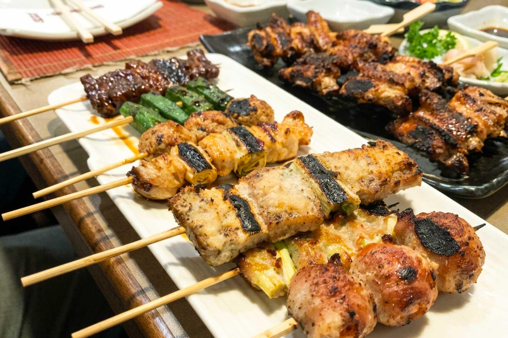 long white ceramic platter filled with freshly grilled Japanese skewers, including seasoned pork belly, chicken and scallion (negima), meatballs, grilled okra, and glazed beef. Background Plate: A black platter holding char-grilled chicken wings (tebasaki) next to small condiment dishes with dipping sauce, pickled ginger, and wasabi on a wooden dining table.