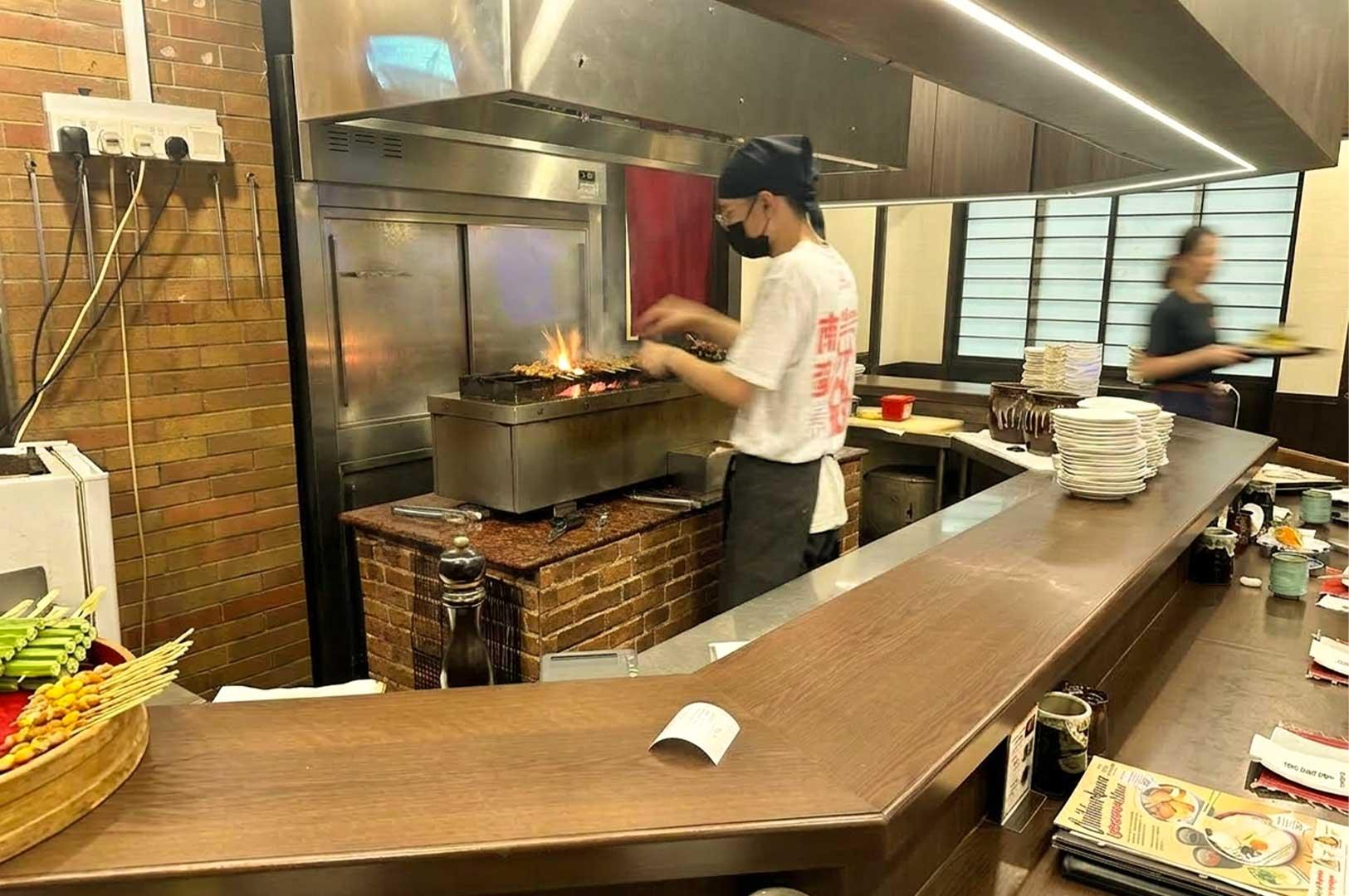 A wooden counter seating area displaying printed menus, ceramic cups, stacked plates, and a wooden bowl filled with uncooked vegetable and meat skewers. Kitchen Area: A chef wearing a bandana, face mask, and white graphic t-shirt grilling yakitori skewers over an open flame on a charcoal grill station built with exposed brick. Background Setting: The warm interior of the Japanese restaurant featuring traditional Shoji sliding doors and a server moving past in the background.