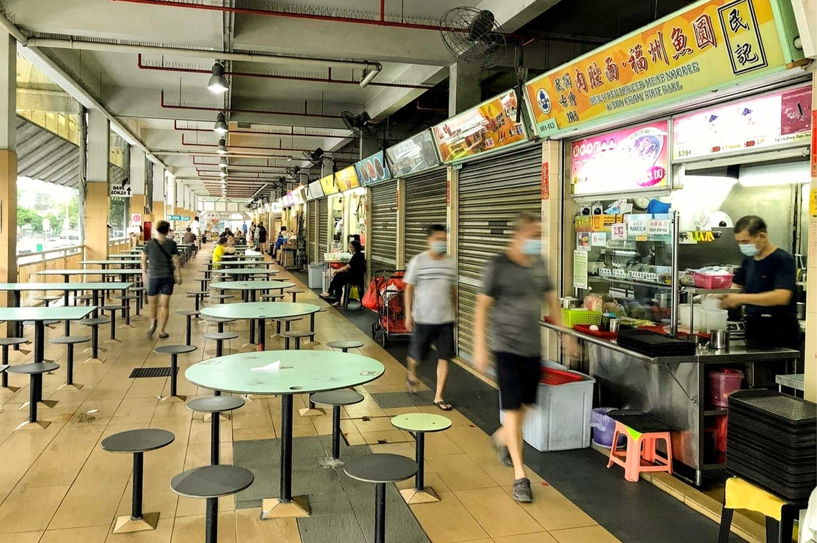 An open-air corridor of a Singaporean hawker center lined with shuttered and active food stalls, featuring a hawker vendor in a face mask preparing food behind a glass-front counter while masked pedestrians walk past fixed circular green tables and grey stools.