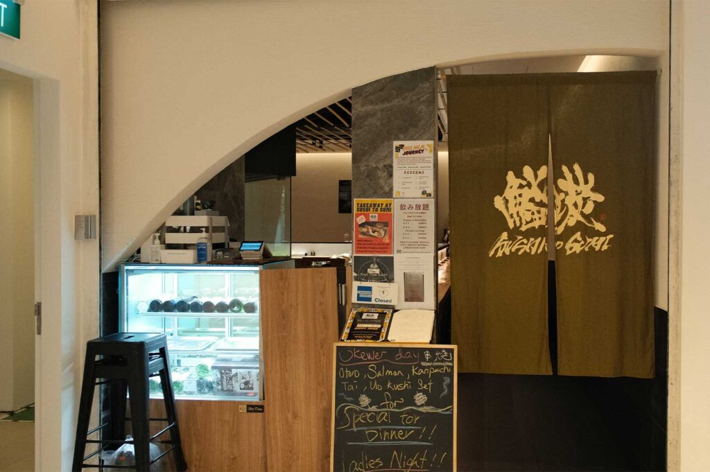 The entrance to the Japanese restaurant "Sushi to Sumi," featuring a dark green noren curtain emblazoned with gold calligraphic text on the right and an arched entryway leading inside on the left, complete with a glass display case, informational posters, and a handwritten chalkboard menu advertising dinner specials at the front.