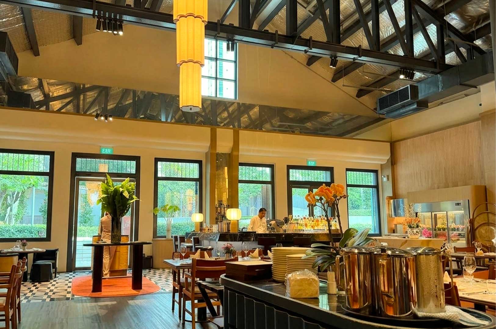 A wide view of a spacious, upscale restaurant interior featuring high ceilings with exposed dark wooden trusses and tall, tiered cylindrical pendant lights hanging down. Sunlight streams through large black-framed windows overlooking lush greenery outside, illuminating neatly set wooden dining tables, a fully stocked bar counter with a bartender in the background, and a serving station in the foreground displaying stacked plates, orchids, and metallic pitchers.