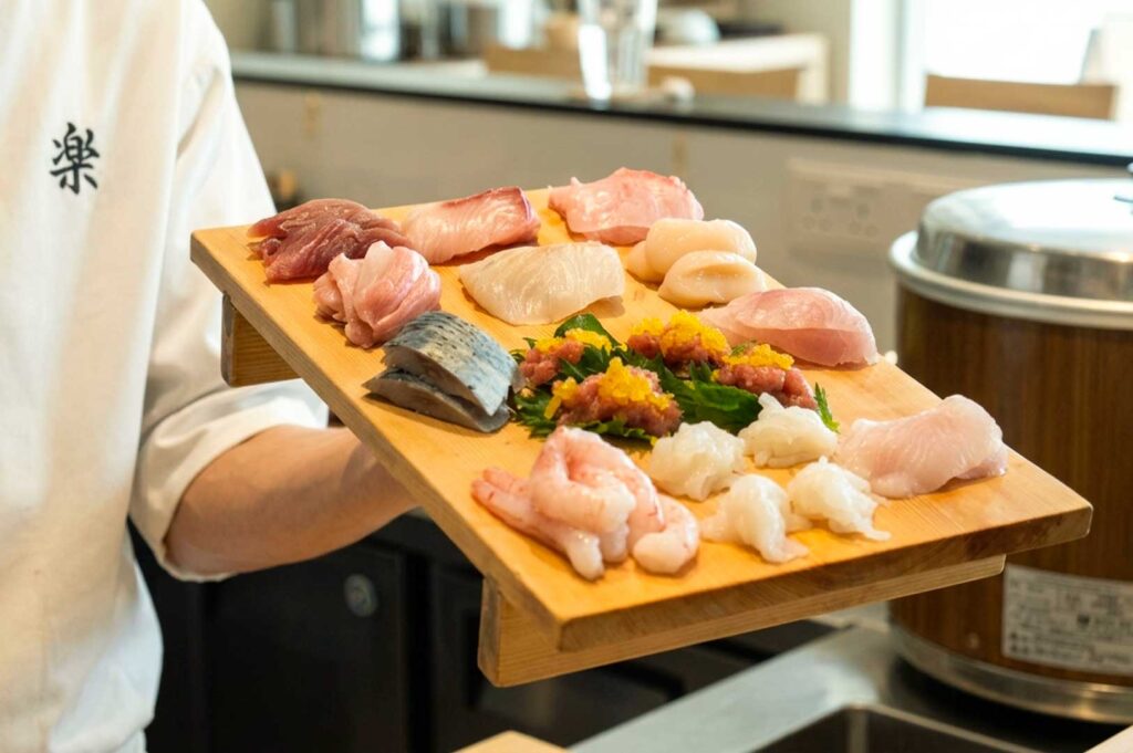 A sushi chef in a white uniform holding a wooden platter laden with an assortment of fresh sashimi, including slices of raw tuna, salmon, scallops, sweet shrimp, and seared fish arranged over green leaves.