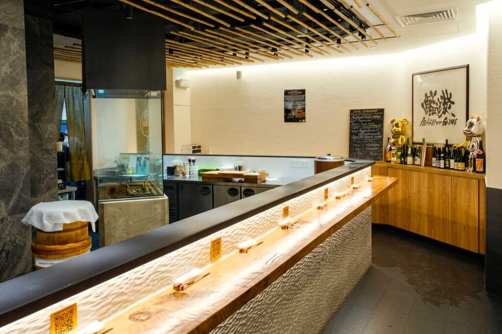 The interior of a modern standing sushi bar featuring an L-shaped illuminated wooden counter set with chopstick rests, a glass-enclosed charcoal grill station on the left, light-colored wooden cabinets with sake bottles on the right, and a ceiling decorated with bamboo poles overhead.