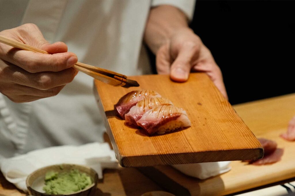 A close-up shot of a sushi chef applying a soy sauce glaze with a small brush onto three pieces of fresh nigiri sushi arranged on a wooden wooden board.