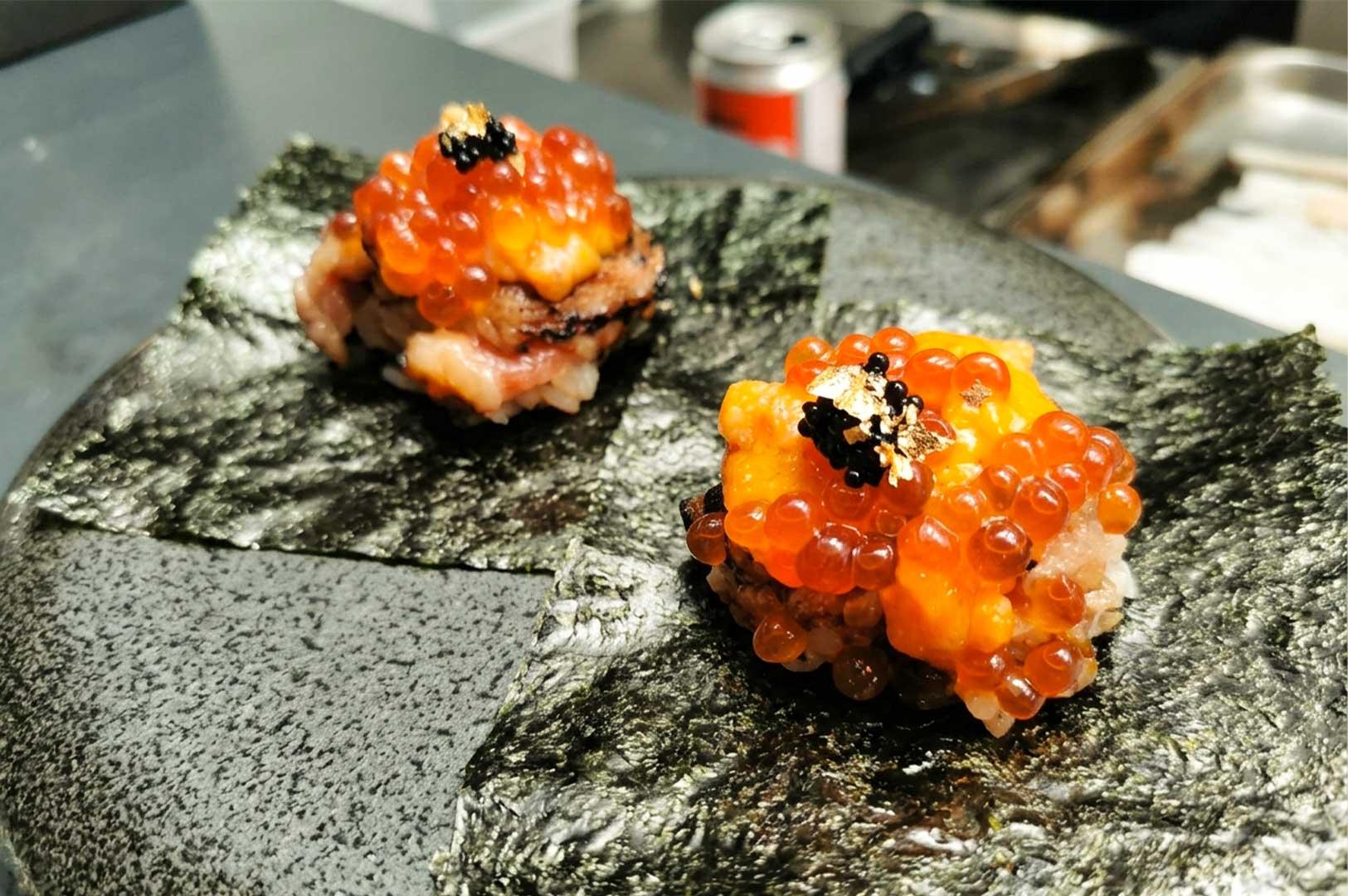 Two open-faced sushi handrolls are presented on a textured black plate, each sitting atop a square sheet of crisp nori seaweed. The handrolls are lavishly layered with mounds of rice, creamy sea urchin (uni), bright orange salmon roe (ikura), a dollop of black caviar, and a delicate speck of edible gold leaf on top.