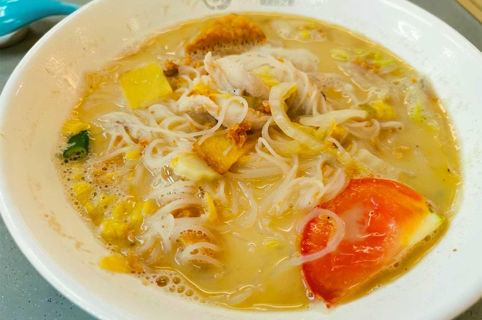 A close-up view of a bowl of sliced fish soup with milky broth and thin rice vermicelli noodles from Jun Yuan House of Fish, garnished with tender white fish slices, a red tomato wedge, cabbage, and crispy fried egg floss.