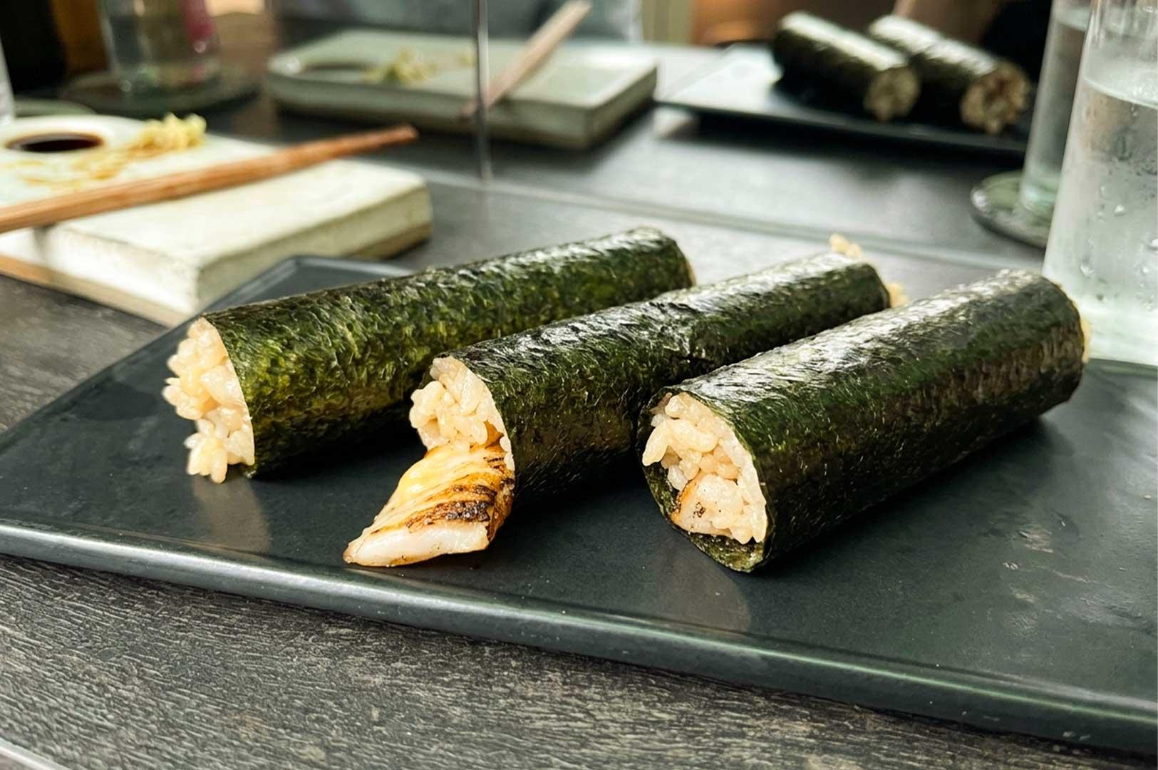 Three cylindrical sushi handrolls wrapped in crisp green nori seaweed are arranged diagonally across a dark grey rectangular plate placed on a table. The handrolls are filled with seasoned brown sushi rice, with one roll in the center featuring a piece of charred, seared fish protruding from the end. In the soft-focused background, additional serving dishes, chopsticks, glasses of water, and another plate of handrolls sit on the dining counter.