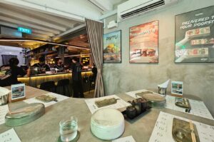 A modern, industrial-style sushi handroll bar features a long concrete counter set with paper placemats, ceramic plates, sauce pots, and chopsticks ready for diners. In the background, staff members in dark jerseys interact behind an illuminated bar, while framed promotional posters, an exposed ceiling structure, and a wall-mounted air conditioner frame the right side of the dining room.