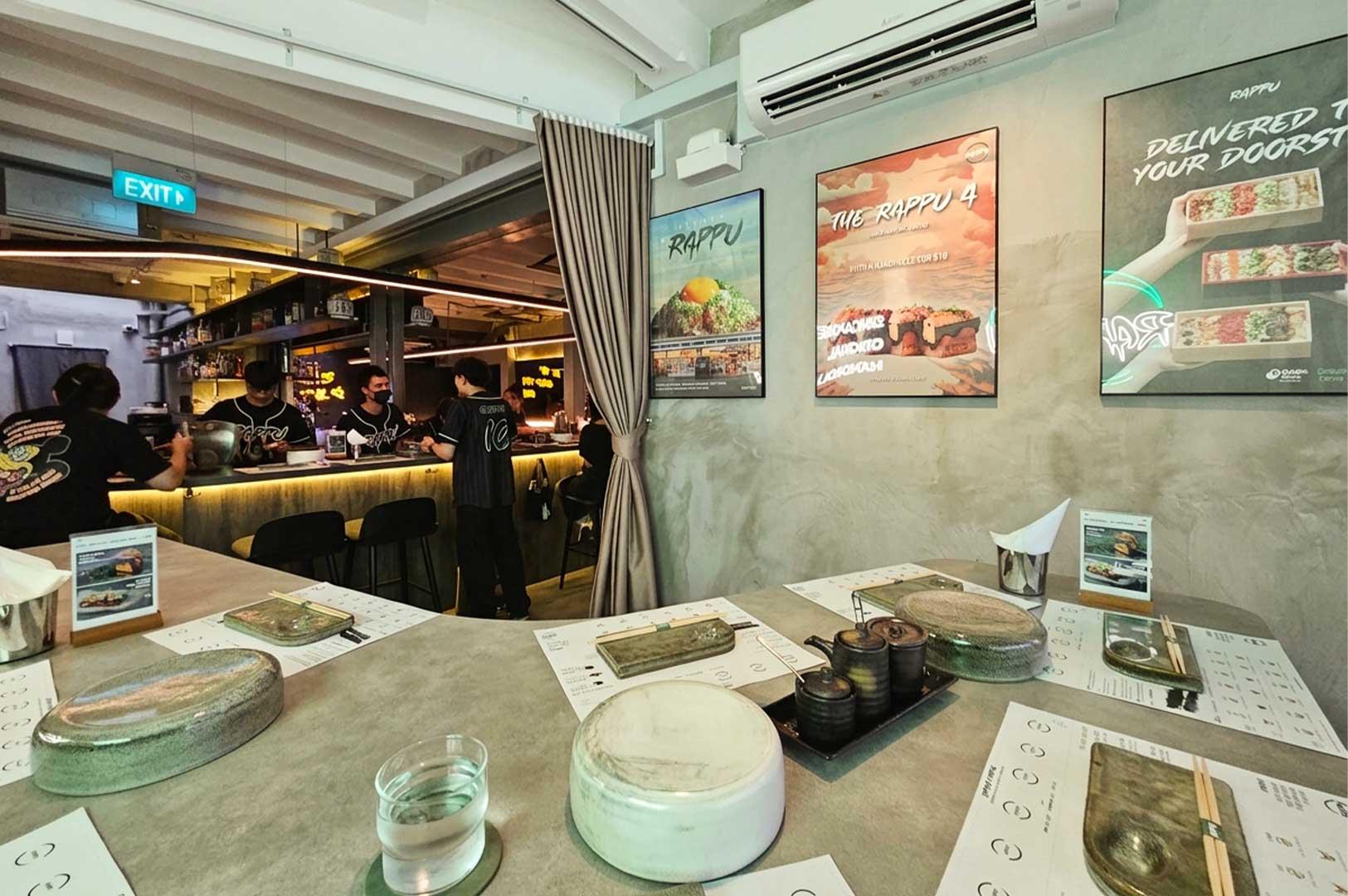 A modern, industrial-style sushi handroll bar features a long concrete counter set with paper placemats, ceramic plates, sauce pots, and chopsticks ready for diners. In the background, staff members in dark jerseys interact behind an illuminated bar, while framed promotional posters, an exposed ceiling structure, and a wall-mounted air conditioner frame the right side of the dining room.