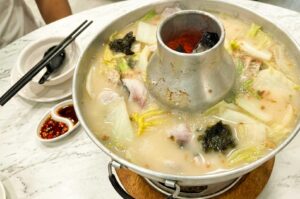 A traditional charcoal-heated metal steamboat pot filled with simmering milky fish head broth, Napa cabbage, seaweed, yam pieces, and fresh fish slices, set on a marble dining table alongside a small dual-compartment saucer with sliced chili and soy sauce, a bowl, and black chopsticks.
