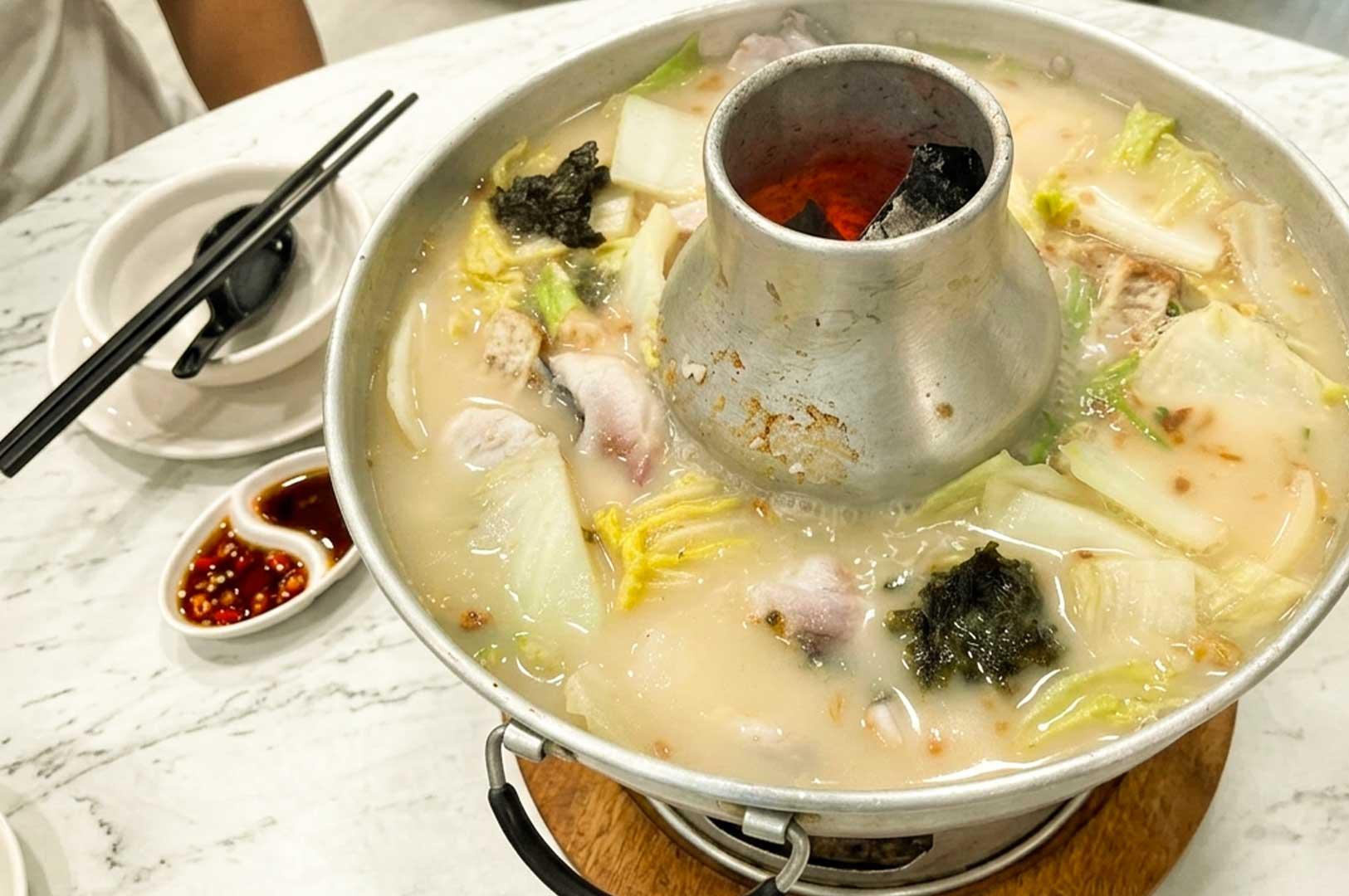 A traditional charcoal-heated metal steamboat pot filled with simmering milky fish head broth, Napa cabbage, seaweed, yam pieces, and fresh fish slices, set on a marble dining table alongside a small dual-compartment saucer with sliced chili and soy sauce, a bowl, and black chopsticks.