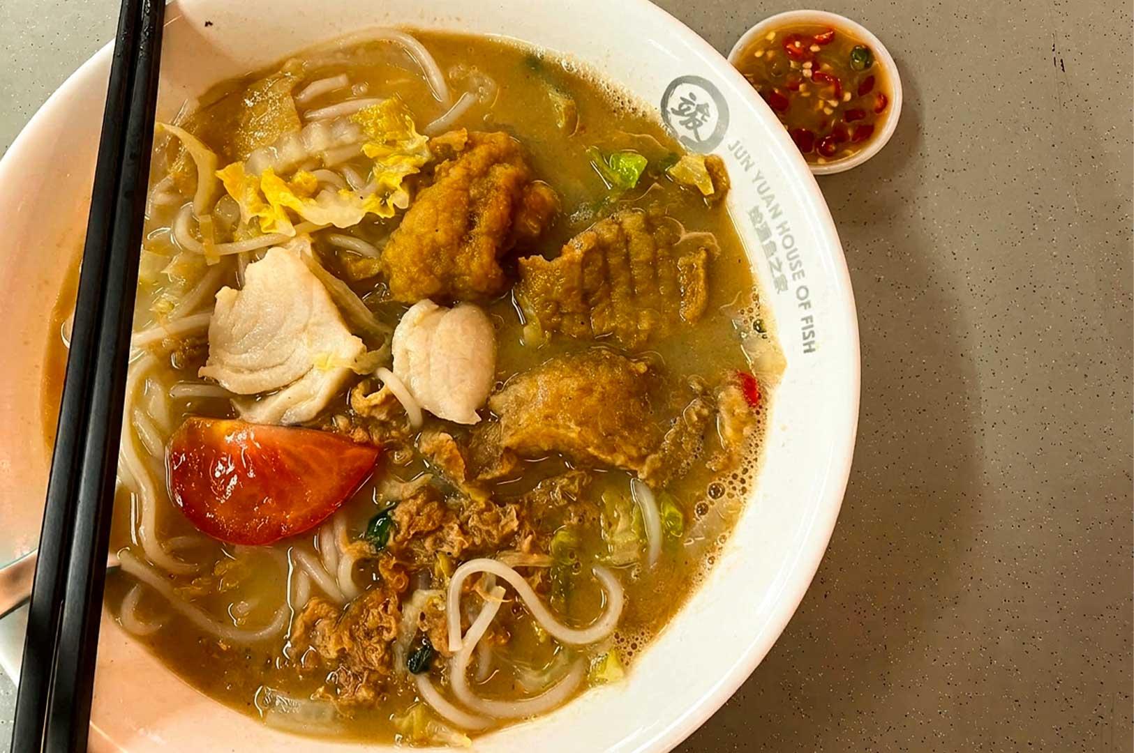 A bowl of fish noodle soup from Jun Yuan House of Fish, filled with rich broth, thick rice vermicelli, slices of fresh fish, deep-fried fish nuggets, a tomato wedge, greens, and crispy fried egg strands, served with a small dish of sliced chili padi in soy sauce on the side.